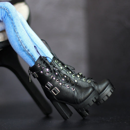 A doll seated on a black chair with one leg extended, wearing black lace-up platform ankle boots with metal studs and buckles, paired with light blue jeans against a dark studio background.